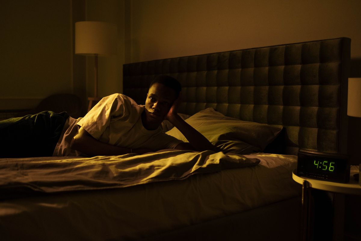 A man lying awake in bed at night, an alarm clock showing 4:56 glowing in the dark