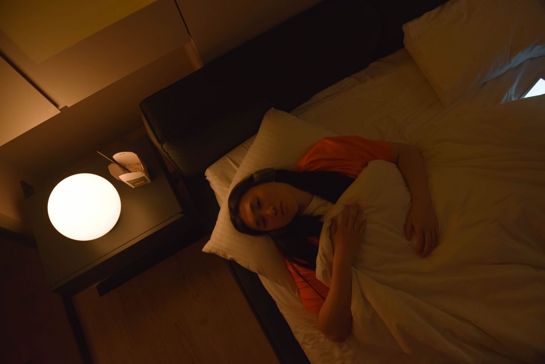 A woman lying awake in bed in a dimly lit room at night, eyes open, warm lamplight beside her